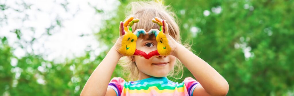 Happy child with colorful painted hands making a heart shape outdoors after passing childcare background checks, ensuring safe supervision.