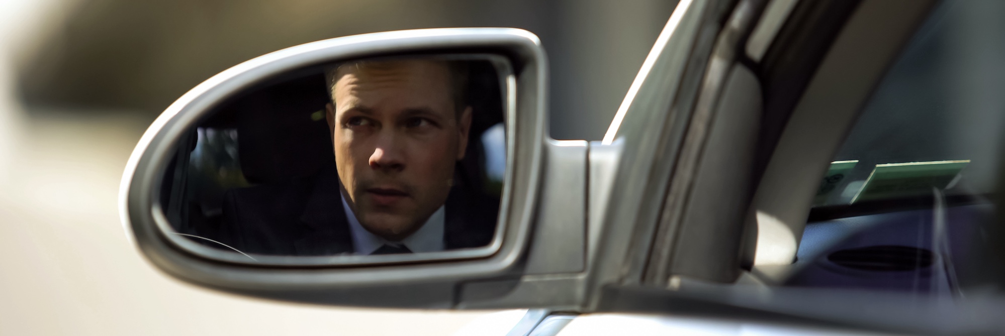 A professional is conducting a due diligence investigation while observing the subject through the vehicle side mirror during surveillance.
