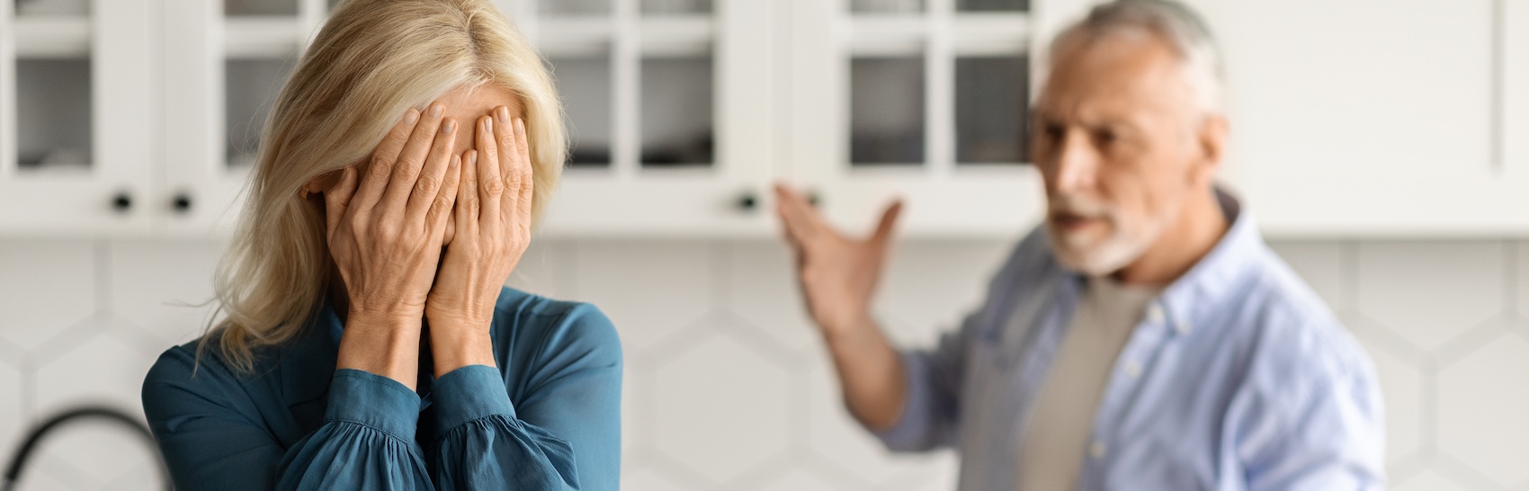 A distressed senior woman covering her face with her hands during elder exploitation investigation while her husband argues in the kitchen background.