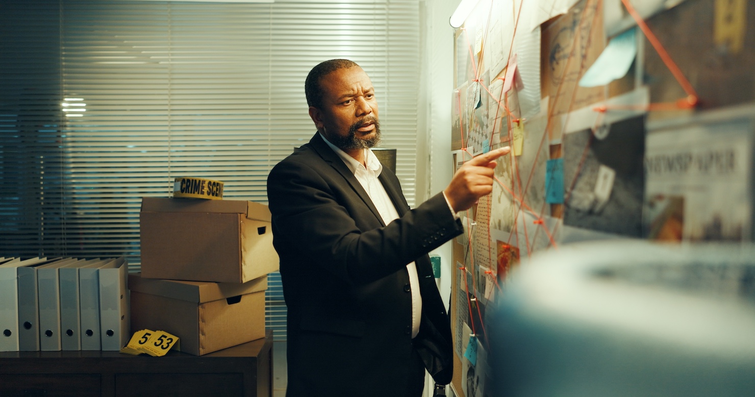 A male private investigator for judgment collection, examining evidence on a wall with red string connections, and crime scene boxes in the investigation office.