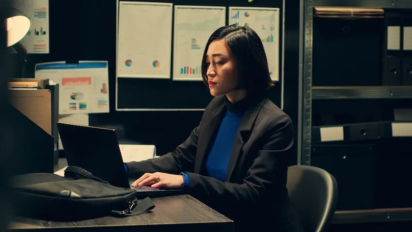 A woman in a business suit works intently on her laptop late at night surrounded by charts and binders depicting the thorough case research an attorney conducts when building a contractor fraud claim on behalf of a client at Upstate Private Investigators in South Carolina.