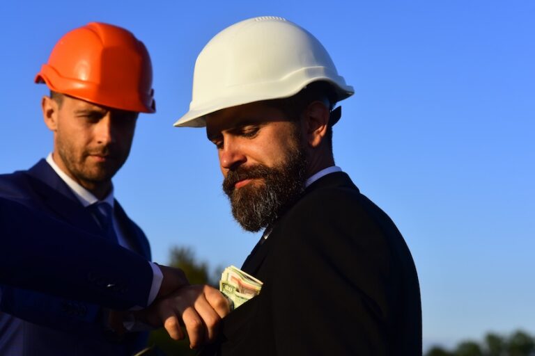 Two men in suits and hard hats exchange cash outdoors under a clear blue sky depicting the kind of underhanded financial exchange that is characteristic of contractor fraud schemes in the construction industry in South Carolina.