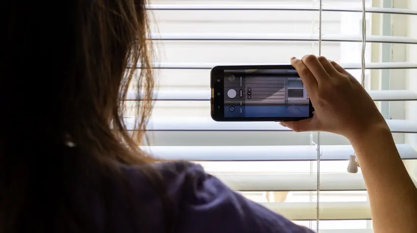 A person peers through window blinds while holding up a smartphone to take a photo capturing the kind of neighbor surveillance and documentation that often serves as evidence in HOA disputes over property violations in South Carolina.