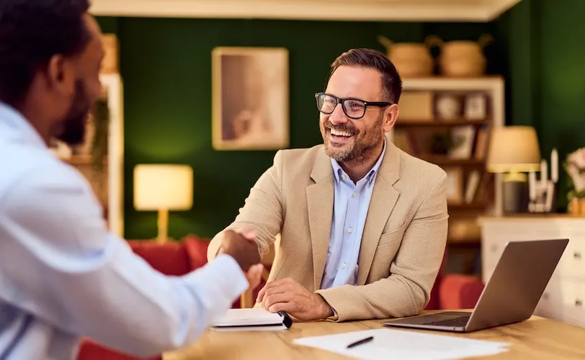 A smiling man in a tan blazer and glasses reaches across a desk to shake hands with a client representing the positive resolution a homeowner can achieve when working with legal counsel to settle HOA disputes at Upstate Private Investigators.
