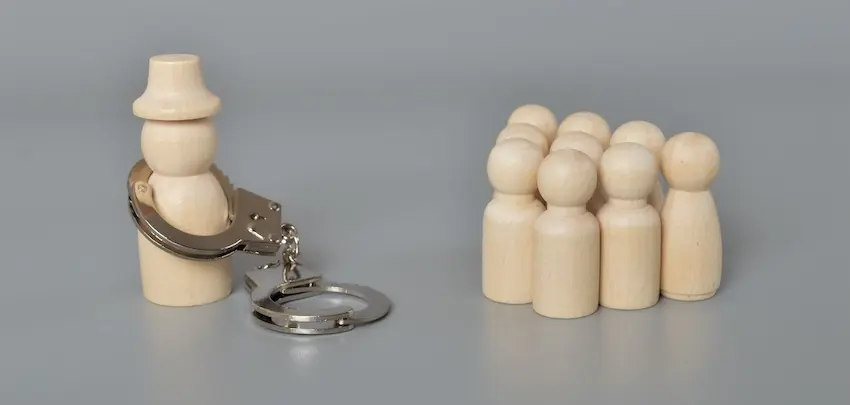 A wooden figure wearing a hat and handcuffs stands separated from a group of plain wooden figures on a gray background symbolizing the arrest and accountability that can result from a contractor fraud investigation in South Carolina.