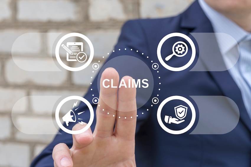 A lawyer from Upstate Private Investigators in a dark navy business suit extends one finger to tap a central dark circular button labeled "CLAIMS" in white capital letters on a glowing digital touchscreen interface floating in front of a white exposed brick wall with four surrounding white circular icons connected by dotted lines representing different aspects of the claims process — a document with a checkmark in the upper left- a magnifying glass with a gear in the upper right- a megaphone with a lightning bolt in the lower left- and a shield with a hand cradling it in the lower right — a visual representation of the comprehensive insurance claims support process from filing and investigation through advocacy and coverage protection in the Upstate of South Carolina.
