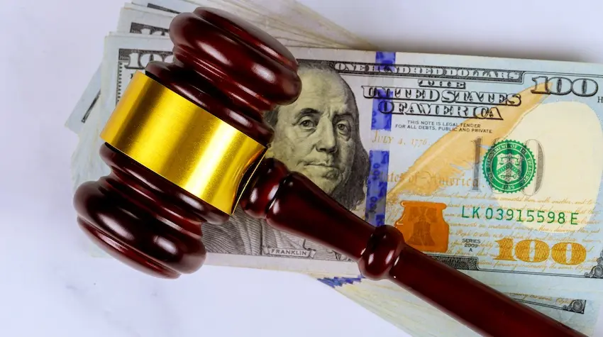 A dark mahogany judge's gavel with a wide shiny gold band lies horizontally across a neatly stacked pile of crisp American hundred-dollar bills on a white marble surface, with Benjamin Franklin's portrait, the words "ONE HUNDRED DOLLARS and UNITED STATES OF AMERICA," a blue security strip- a green treasury seal- and the serial number LK 03915598 E all clearly visible on the top bill beneath the gavel — a direct visual representation of the financial compensation injury victims can recover with proper insurance claims support and effective legal representation from Upstate Private Investigators in the Upstate of South Carolina.