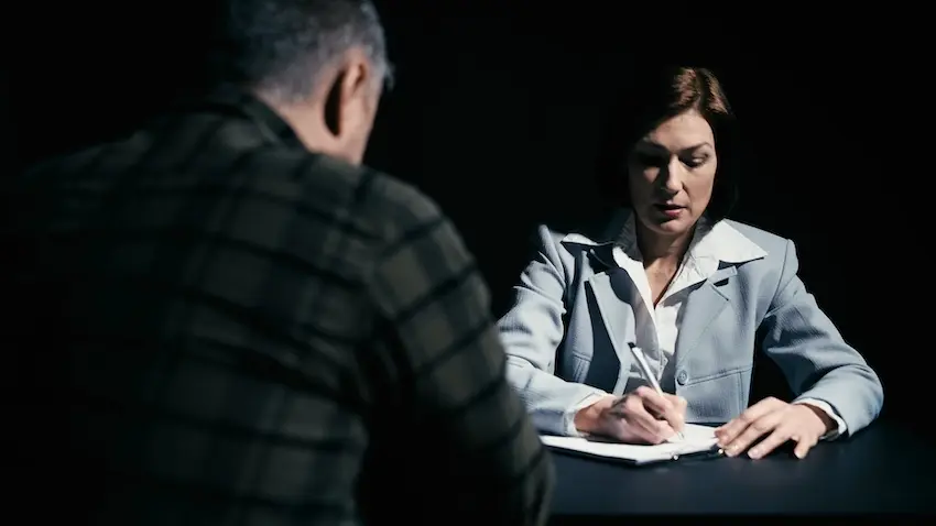A woman with dark hair pulled back- wearing a light gray blazer over a white collared shirt- sits across a dark table in a dimly lit room with a stark black background- leaning slightly forward and writing on a notepad with a pen while speaking directly to a male subject seated across from her whose back is to the camera- visible only from behind in a dark plaid shirt — depicting an intense formal interview or recorded statement session conducted as part of an insurance claims support investigation following an accident in the upstate of South Carolina.